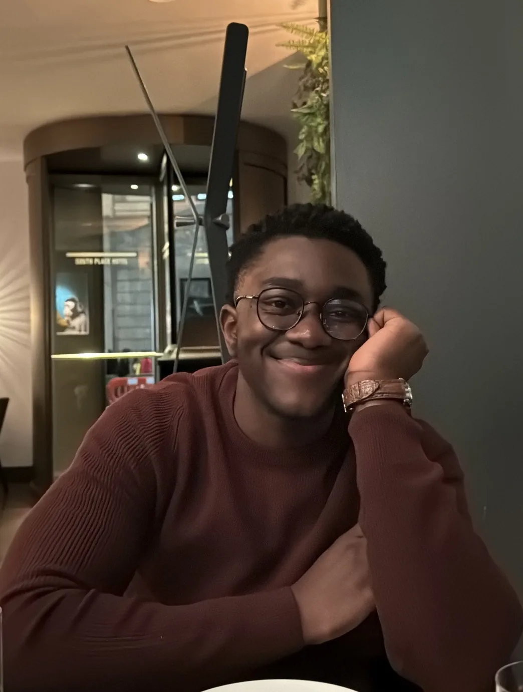 Yooku Obiri, in glasses and a burgundy jumper, smiling and resting his head on his hand.